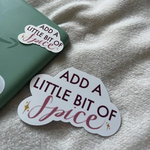 Puede incluir: Pegatina blanca en forma de nube con el texto "Add a little bit of spice" en letras granate y rosa. La pegatina tiene un acento de purpurina dorada.