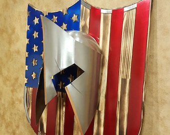 May include: Wooden shield featuring the American flag design with red and white stripes, blue stars, and a silver Spartan helmet. The shield has a rustic, burnt wood appearance with a metallic trim.