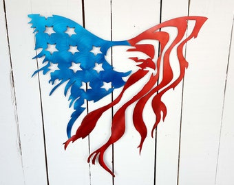 May include: A metal wall art piece depicting a stylized American flag. The flag features a blue section with white stars and red and white stripes, with a distressed, flowing design. The artwork is mounted on a white wooden background.