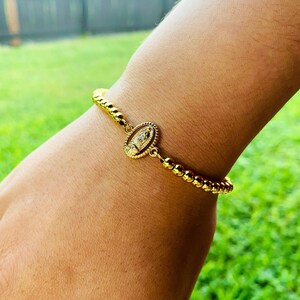 May include: A gold bracelet with a small oval charm featuring a religious image. The bracelet is made of small gold beads.