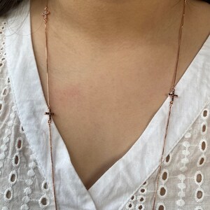 May include: A long rose gold chain necklace with small black cross charms. The necklace is worn by a person with a white shirt.