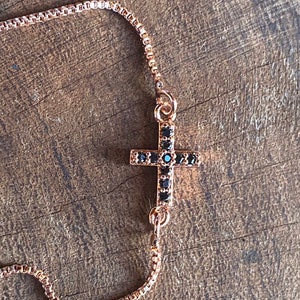 May include: A gold chain bracelet with a black cross charm. The cross is adorned with small black stones.