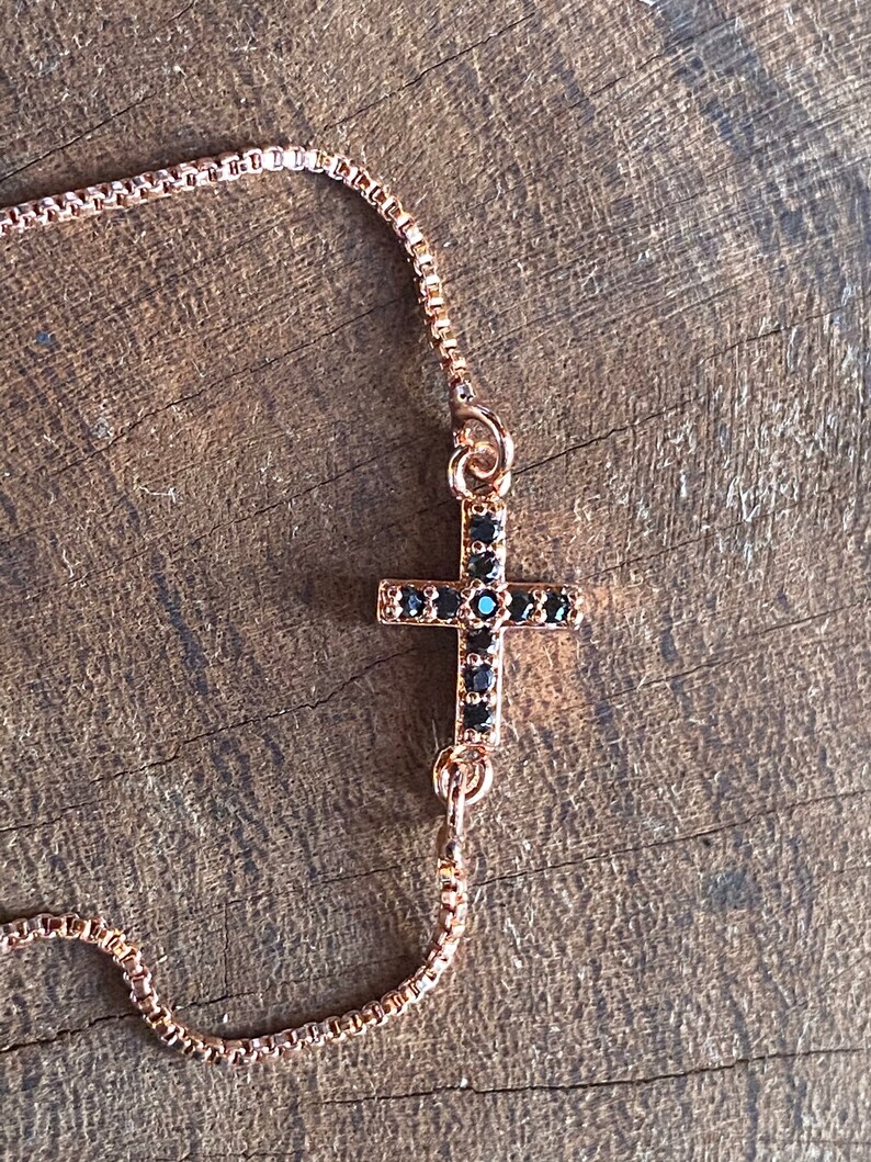 May include: A gold chain bracelet with a black cross charm. The cross is adorned with small black stones.