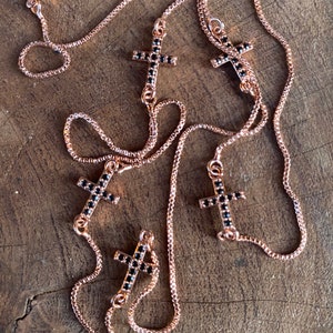 May include: Multiple rose gold chains with black crystal cross pendants. The chains are layered and the crosses are of varying sizes.