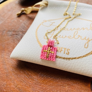 May include: A gold chain necklace with a pink and gold beaded pendant in the shape of a gift box. The pendant has a gold cross design. The necklace is displayed on a white pouch with gold text.