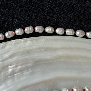 Può includere: Primo piano di una collana di perle con perle bianche di forma ovale infilate su un cordoncino rosso. La collana è appoggiata su una superficie bianca e iridescente, con uno sfondo scuro e strutturato.