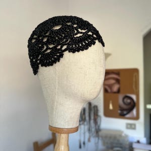Può includere: Un copricapo all'uncinetto nero caratterizzato da un intricato motivo in pizzo traforato. L'accessorio è aderente alla parte superiore di una testa di manichino beige e presenta un bordo smerlato che segue il contorno della fronte.