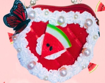 May include: A red heart-shaped purse with a white trim and a watermelon slice design. The purse has a pearl chain strap and a butterfly charm. The text "PICNIC PRINCESS" is printed in green letters on a pink background.