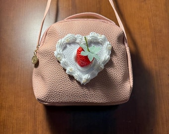 May include: A pink crossbody bag with a textured surface. The bag features a heart-shaped embellishment with a red strawberry and white decorative accents. The bag has a top handle and a long strap.