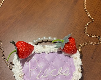May include: A light purple purse with a gold chain strap, featuring a quilted design and the word "Pisces" in cursive. The purse is decorated with faux strawberries and white pearl accents.