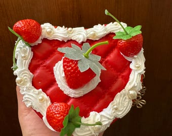 May include: A red, heart-shaped purse with a quilted design, adorned with white frosting-like trim and faux strawberries. The purse features pearl-like embellishments and a gold chain strap.
