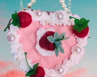May include: A heart-shaped pink handbag with a quilted design, adorned with faux strawberries, pearls, and white frosting-like trim. The bag has a pearl handle and chain straps. The text "STRAWBERRY DREAM" is at the bottom.