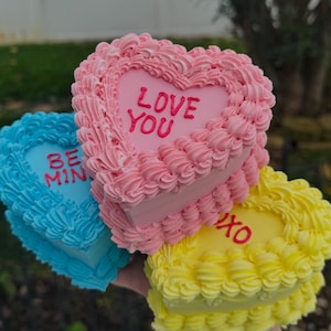 May include: Three heart-shaped cakes in pink, blue, and yellow, each with a different message. The pink cake says "LOVE YOU", the blue cake says "BE MINE", and the yellow cake says "XO". The cakes have a decorative frosting.