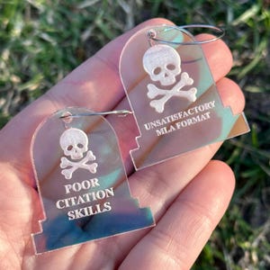 May include: A pair of iridescent acrylic earrings shaped like tombstones. The earrings feature a skull and crossbones design with the text "Unsatisfactory MLA Format" and "Poor Citation Skills".