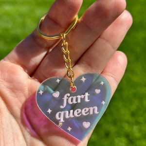 May include: A heart-shaped iridescent keychain with a gold chain and a white inscription that reads "fart queen".