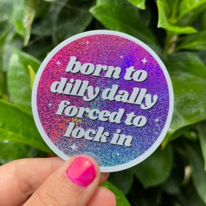 Könnte beinhalten: Ein runder Aufkleber mit holografischem Finish. Der Aufkleber hat einen rosa-violetten Farbverlauf mit weißem Text, der "born to dilly dally forced to lock in" lautet.
