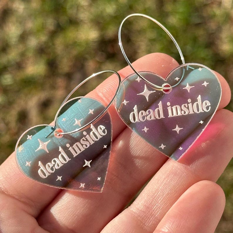 May include: A pair of iridescent heart-shaped earrings with silver hoops. The hearts have the text "dead inside" printed on them.