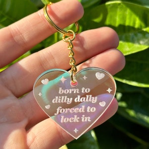May include: An iridescent heart-shaped keyring with a gold chain and the text "born to dilly dally forced to lock in".