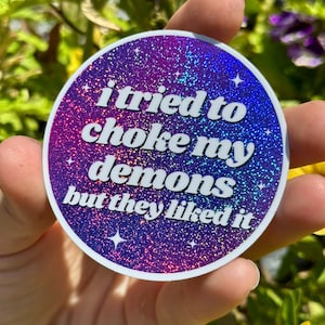 May include: A round sticker with a holographic finish features the text "i tried to choke my demons but they liked it" in white letters. The background is a gradient of purple and blue with glitter.