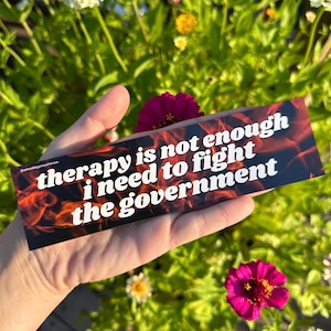 May include: A rectangular bumper sticker with the text "therapy is not enough I need to fight the government" in white font. The background features a fiery red and orange design. The sticker is held in a hand against a backdrop of green foliage and pink flowers.