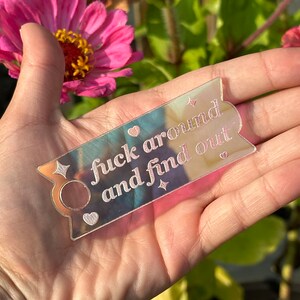 May include: A clear, iridescent acrylic bookmark with the phrase "fuck around and find out" in a cursive font. The bookmark has a banner shape with a small circle and heart design. Decorative star shapes are also included.
