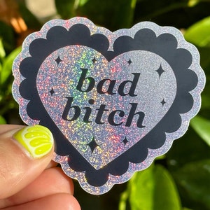 Puede incluir: Una pegatina en forma de corazón con un borde negro festoneado y un interior plateado brillante. Las palabras "bad bitch" están impresas en escritura negra. Cuatro pequeñas formas de estrella negra están dispersas por el fondo brillante.