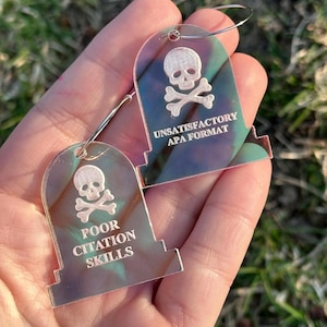 May include: Two iridescent acrylic earrings shaped like tombstones with a skull and crossbones design. The text on the earrings reads "Unsatisfactory APA Format" and "Poor Citation Skills".