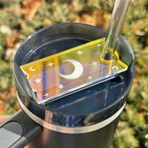 May include: A black travel mug with a yellow and iridescent moon and stars design on the lid. The lid has a hole for a straw, and a straw is inserted through the hole.