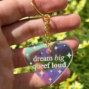 May include: A iridescent heart-shaped keychain with a gold chain. The keychain has the text "dream big queef loud" written on it.