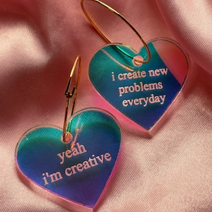 May include: A pair of iridescent heart-shaped earrings with gold hoops. The left earring says "yeah i'm creative" and the right earring says "i create new problems everyday".
