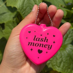 May include: A bright neon pink heart-shaped plastic container featuring the black text "lash money" printed in the centre, surrounded by small black heart and star icons, attached to a silver-toned metal ball chain for hanging.