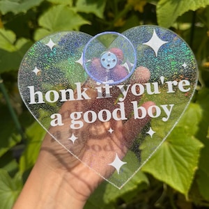 May include: A heart-shaped clear acrylic sign with a holographic glitter finish and a suction cup attached to the top. The sign features white text that reads "honk if you're a good boy" and is decorated with several four-pointed star icons.