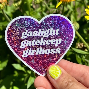 Peut inclure: Un autocollant en forme de cœur avec un fond violet pailleté et un texte blanc qui dit "gaslight gatekeep girlboss".