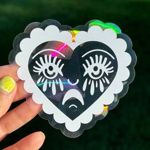 May include: A black and white heart-shaped sticker with a crying face. The heart has a white outline and a scalloped edge. The sticker is on a holographic background.