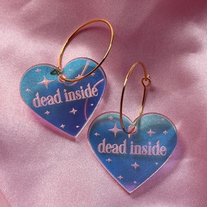 May include: A pair of gold hoop earrings with iridescent blue heart-shaped charms. The charms have the text "dead inside" printed on them with white stars.