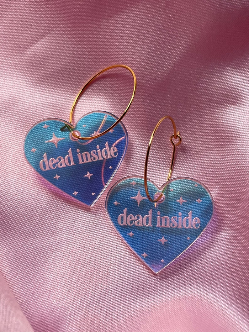May include: A pair of gold hoop earrings with iridescent blue heart-shaped charms. The charms have the text "dead inside" printed on them with white stars.