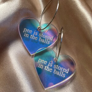 May include: A pair of heart-shaped earrings with iridescent surfaces. Each earring has the phrase "pee is stored in the balls" etched in white. The earrings are suspended from silver-colored hoops.