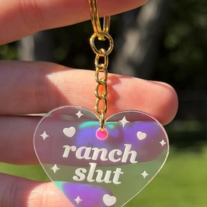 May include: A heart-shaped acrylic keychain with an iridescent finish, featuring the white text "ranch slut" printed in the center. The design includes small white heart and star icons. It is attached to a gold-toned metal split ring and chain.
