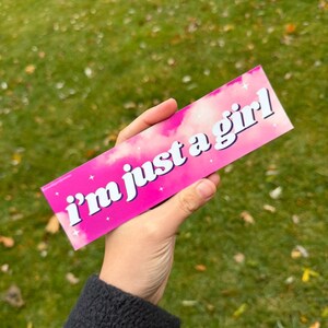 May include: A pink rectangular sticker with the text "i'm just a girl" in white. The sticker has a cloudy design with small white stars. The sticker is held in a hand against a green grassy background.