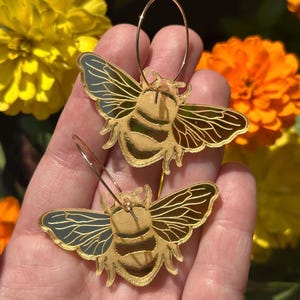 May include: A pair of gold-colored bee-shaped earrings with intricate wing detailing. The earrings are suspended from thin, circular hoops. The background features yellow and orange flowers, creating a vibrant contrast.
