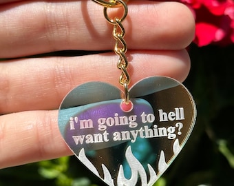 May include: A heart-shaped keychain with a gold chain and a white flame design. The keychain is made of clear acrylic and has the text "i'm going to hell want anything?" printed on it.