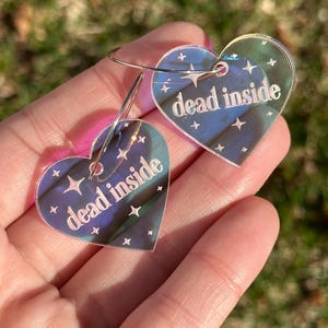 May include: A pair of iridescent heart-shaped earrings with the text "dead inside" on each. The earrings have a silver hoop and are decorated with small white stars.