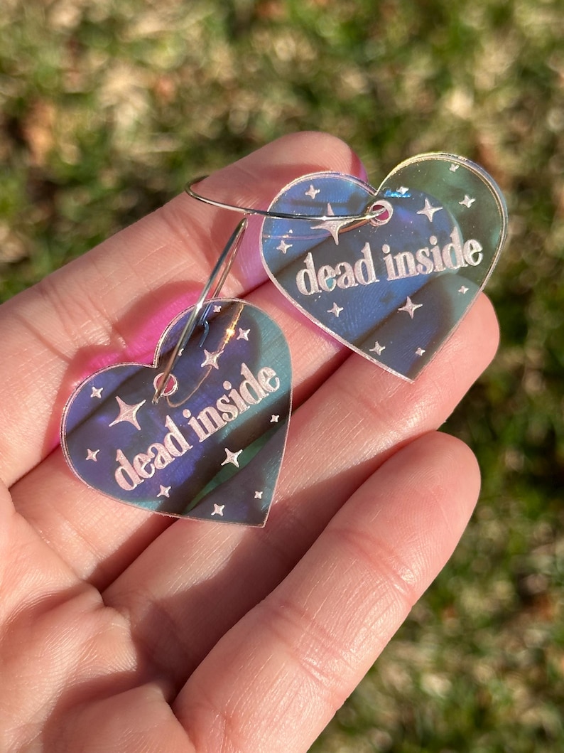 May include: A pair of iridescent heart-shaped earrings with the text "dead inside" on each. The earrings have a silver hoop and are decorated with small white stars.