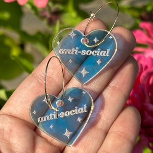 May include: A pair of gold hoop earrings with clear heart-shaped charms. The charms have a glittery finish and the text "anti-social" written in white.