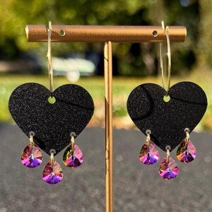 Könnte beinhalten: Ein Paar schwarze herzförmige Ohrringe mit goldenen Reifen. Jeder Ohrring hat drei schillernde, tropfenförmige Kristalle, die von unten herabhängen. Die Ohrringe werden auf einem goldenen Ständer vor einem verschwommenen grünen Hintergrund präsentiert.