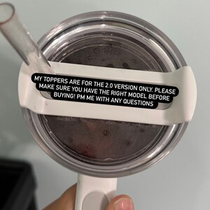 May include: A clear, round tumbler lid with a white handle and straw. The lid has a white plastic holder for the straw. Text on the lid reads: "MY TOPPERS ARE FOR THE 2.0 VERSION ONLY. PLEASE MAKE SURE YOU HAVE THE RIGHT MODEL BEFORE BUYING! PM ME WITH ANY QUESTIONS."