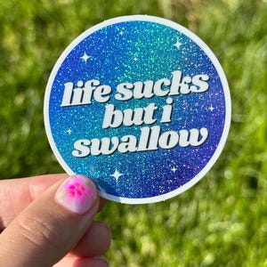 May include: A round blue and green glitter sticker with the text "life sucks but i swallow" in white.