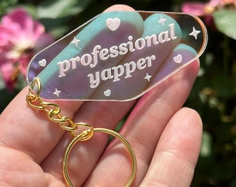 May include: A clear iridescent keychain with a gold chain and ring. The keychain is shaped like a motel sign and has the text "professional yapper" in white lettering.
