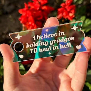 May include: A holographic iridescent keychain with the text "i believe in holding grudges i'll heal in hell" in white lettering. The keychain has a small hole for attaching to a keyring.
