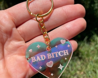 May include: A gold key chain with a heart-shaped charm that says "BAD BITCH" in pink lettering. The charm is iridescent and has a holographic effect.